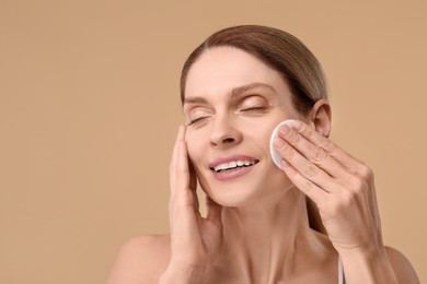 Beautiful woman removing makeup with cotton pad on beige background, space for text