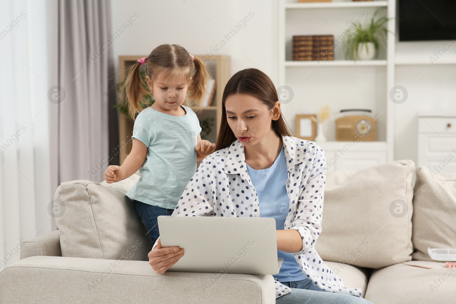 Photo of Woman working remotely at home. Little daughter bothering her mother on sofa in living room