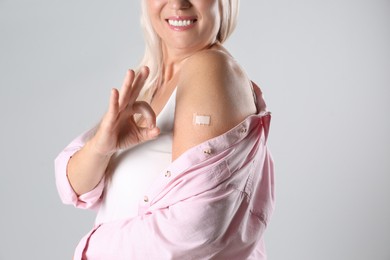 Photo of Happy mature woman showing arm with bandage after vaccination and okay gesture on light background, closeup