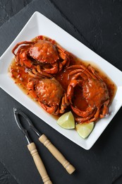 Photo of Delicious boiled crabs with sauce and lime served on black textured table, flat lay