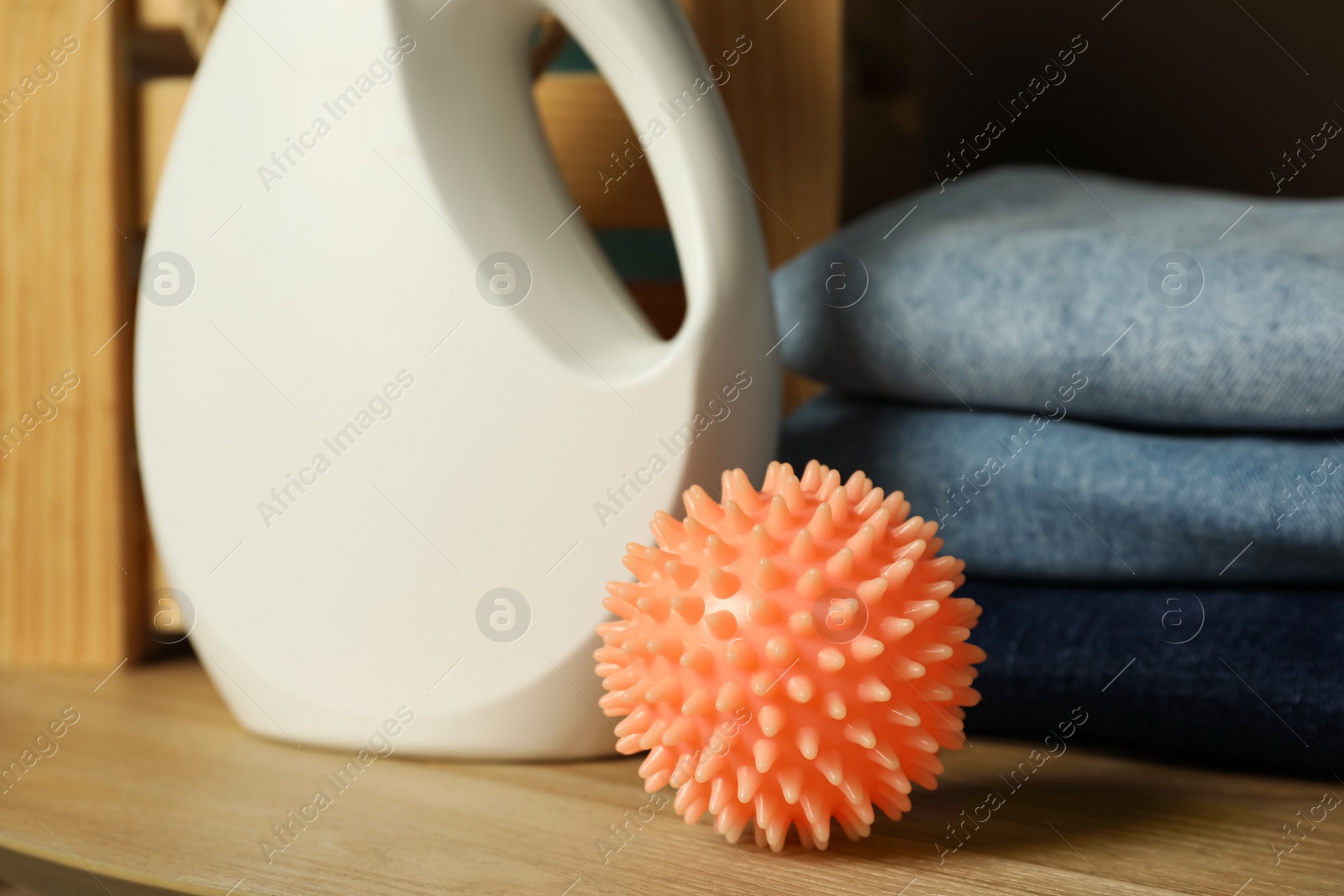Photo of Orange dryer ball, stacked clean clothes and laundry detergent on wooden table, closeup