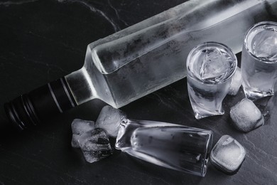 Photo of Bottle of vodka and shot glasses with ice on black table, above view
