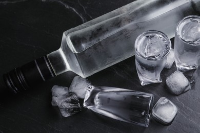 Bottle of vodka and shot glasses with ice on black table, above view