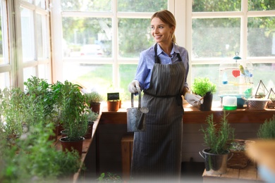 Photo of Young woman with watering can taking care of home plants in shop, view through window