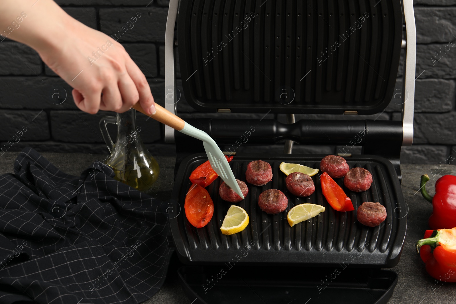 Photo of Woman cooking meat balls with bell peppers and lemon on electric grill at grey table, closeup