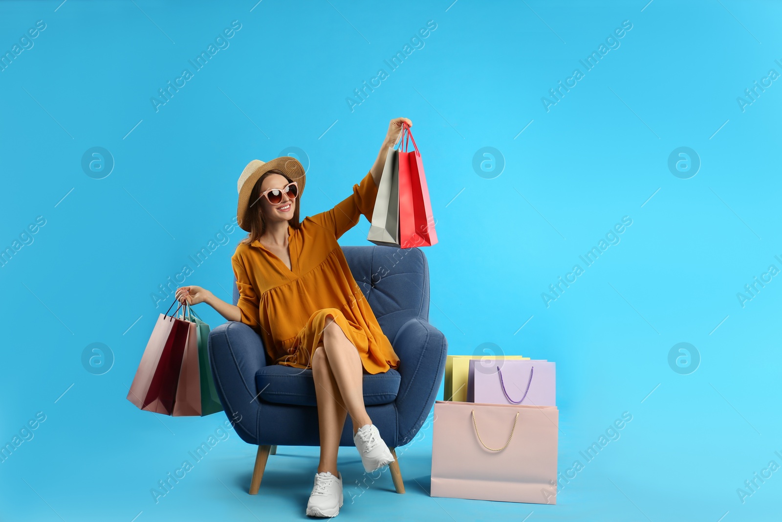 Photo of Beautiful young woman with paper shopping bags in armchair on light blue background