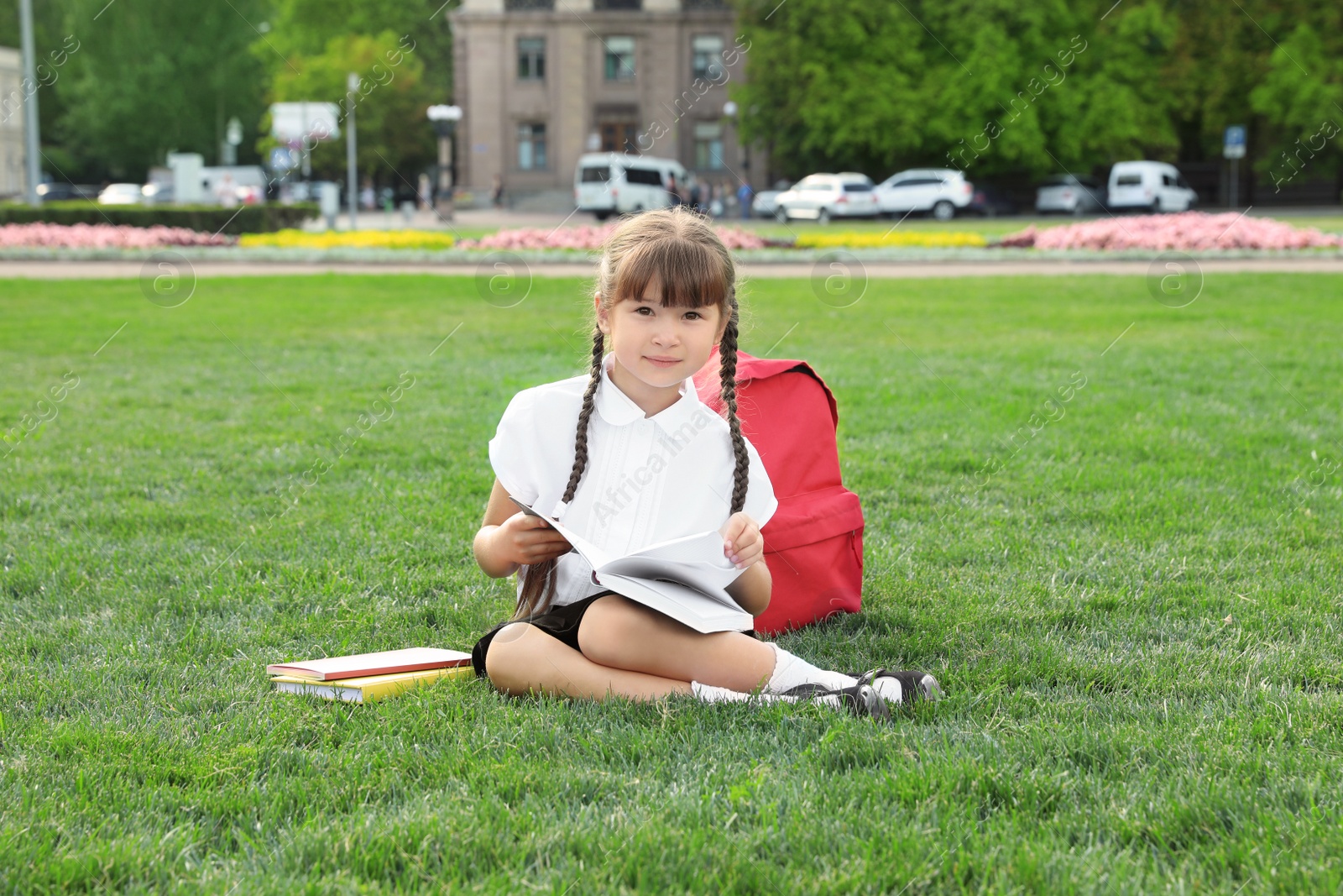 Photo of Cute girl with school stationery reading book on green lawn outdoors