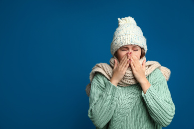 Image of Young woman wearing hat and scarf sneezing on blue background, space for text. Cold symptoms