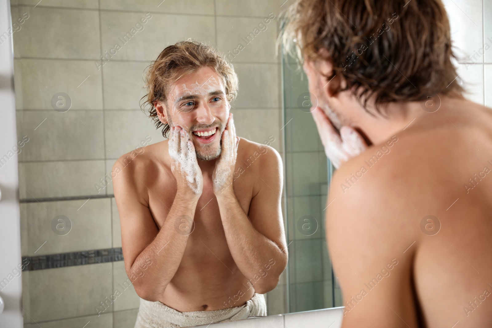 Photo of Young man washing face with soap near mirror in bathroom