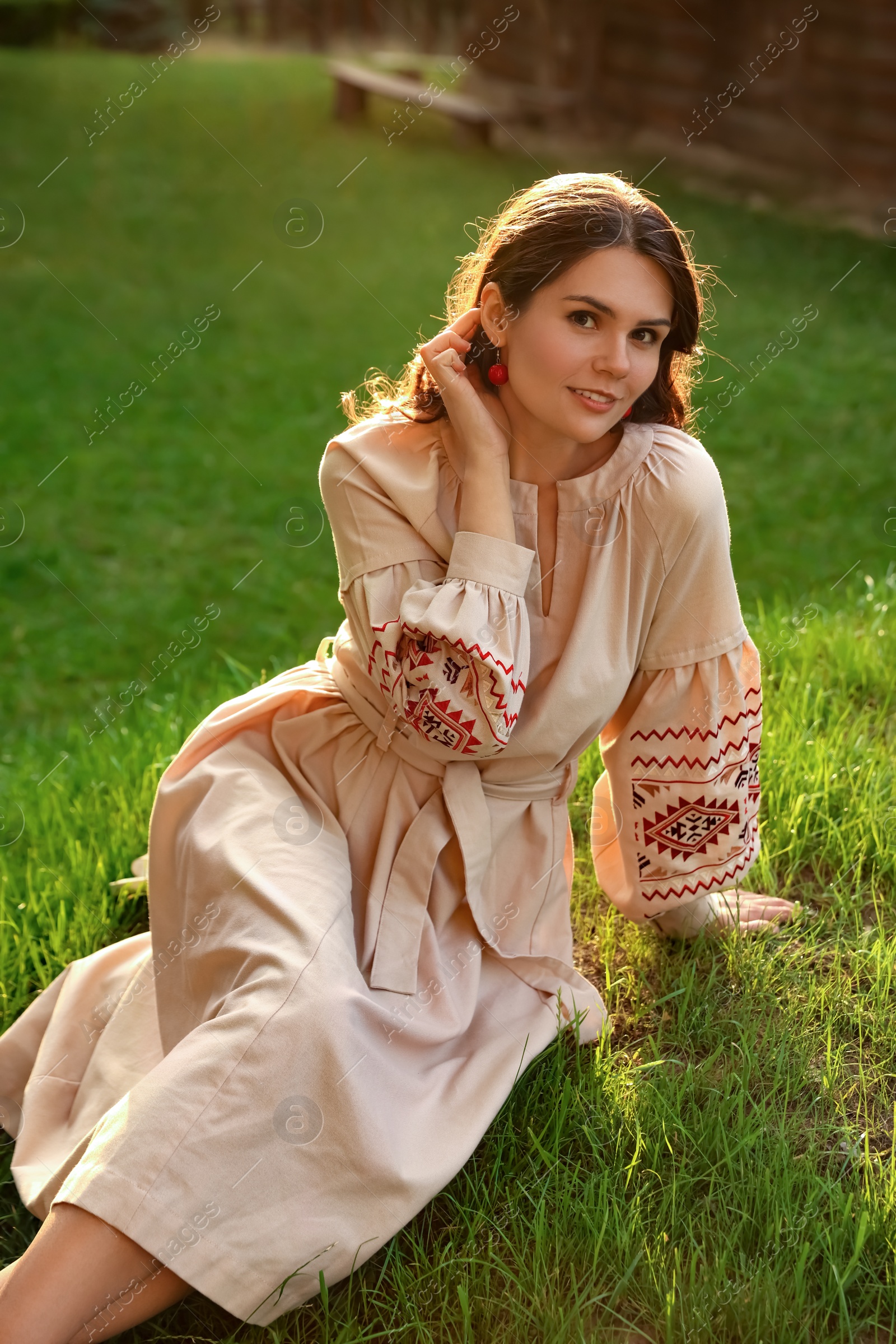 Photo of Beautiful woman in embroidered dress sitting on green grass outdoors. Ukrainian national clothes