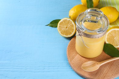 Delicious lemon curd in glass jar, fresh citrus fruits, spoon and green leaves on light blue wooden table, space for text
