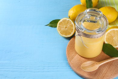 Photo of Delicious lemon curd in glass jar, fresh citrus fruits, spoon and green leaves on light blue wooden table, space for text