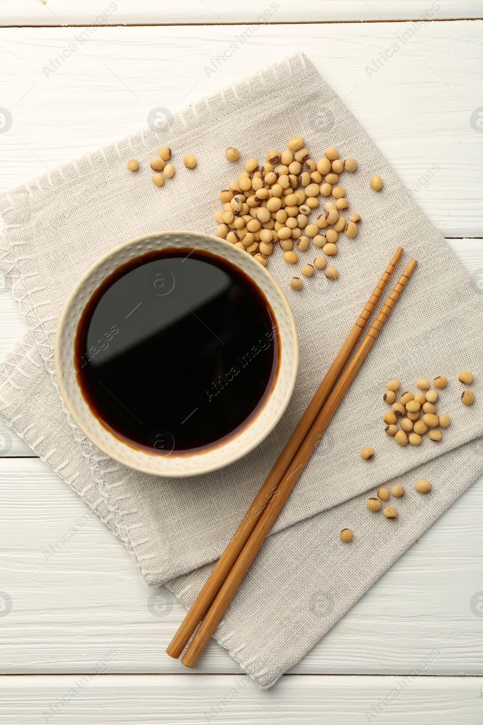 Photo of Tasty soy sauce in bowl, chopsticks and soybeans on white wooden table, top view
