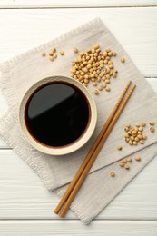 Tasty soy sauce in bowl, chopsticks and soybeans on white wooden table, top view