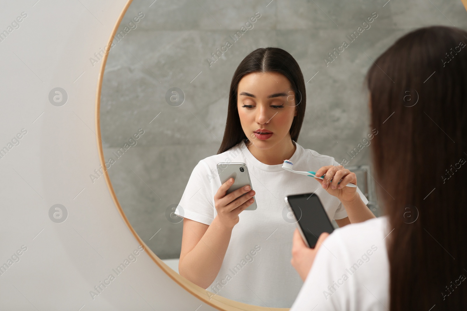 Photo of Beautiful young woman using smartphone while brushing teeth in bathroom. Internet addiction