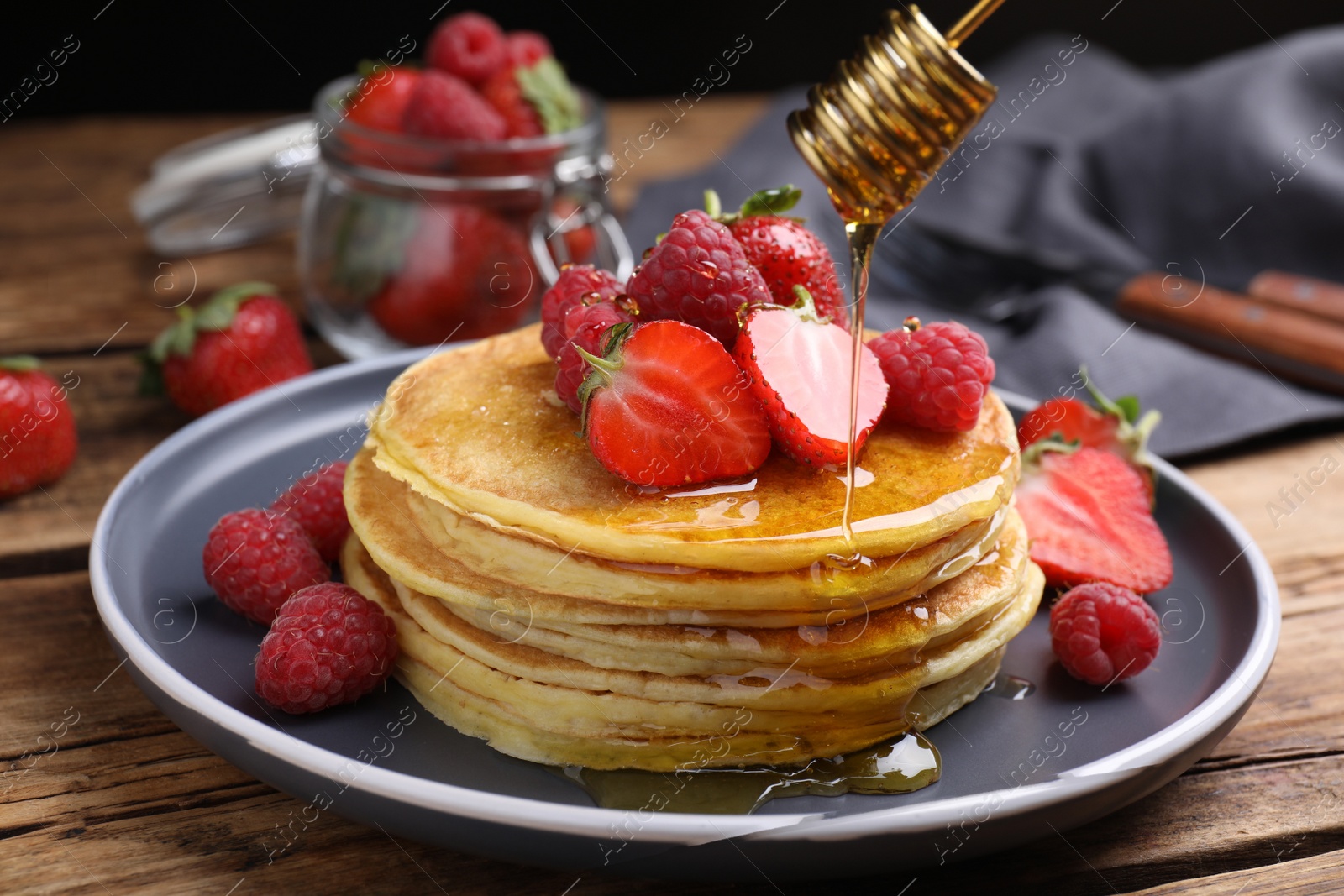 Photo of Pouring honey onto tasty pancakes with fresh berries on wooden table, closeup