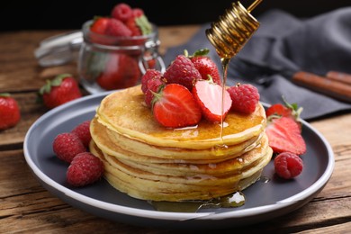 Photo of Pouring honey onto tasty pancakes with fresh berries on wooden table, closeup