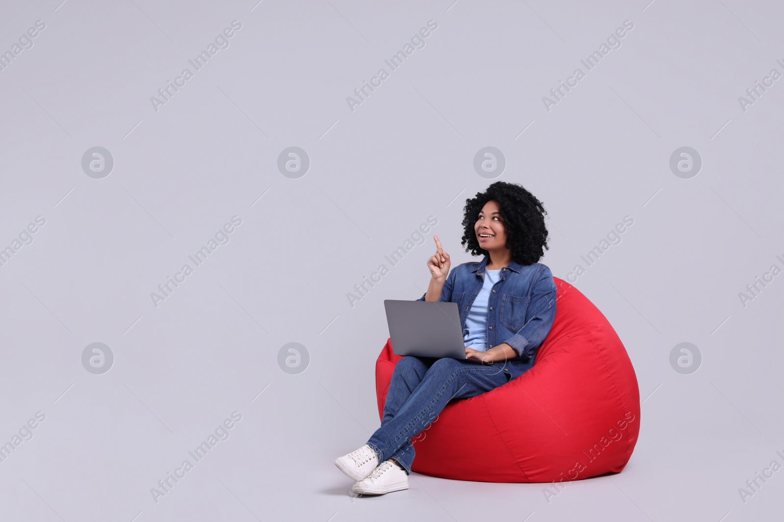 Photo of Happy young woman with laptop pointing at something on beanbag chair against light grey background. Space for text