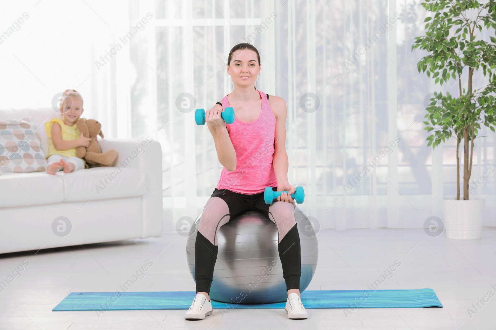 Photo of Woman doing fitness exercises while her daughter sitting on sofa at home