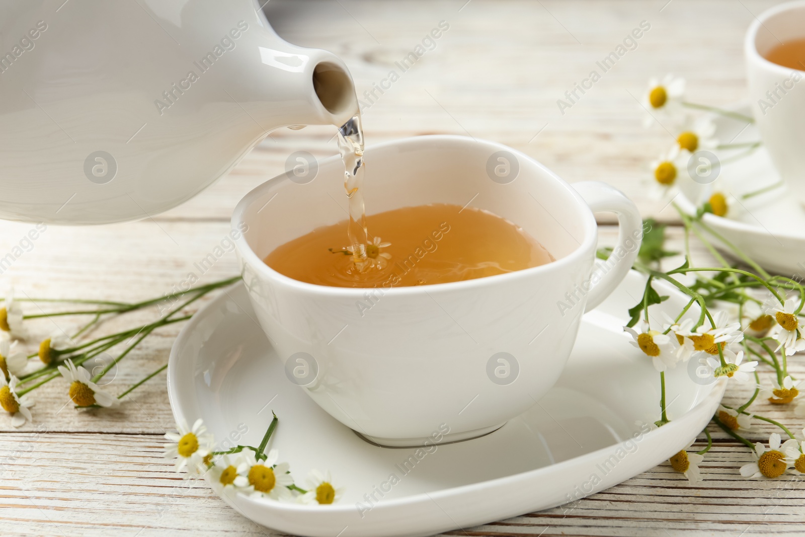 Photo of Pouring tasty chamomile tea into cup on white wooden table