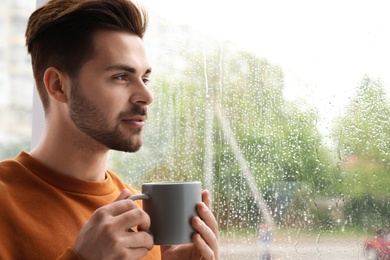 Photo of Thoughtful handsome man with cup of coffee near window indoors on rainy day