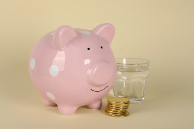 Photo of Water scarcity concept. Piggy bank, coins and glass of drink on beige background