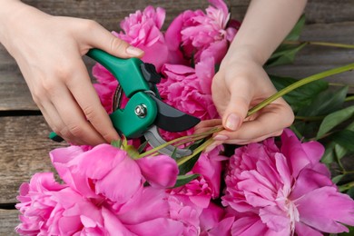 Photo of Woman trimming beautiful pink peonies with secateurs at wooden table, closeup