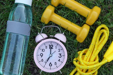 Photo of Alarm clock, skipping rope, dumbbells and bottle of water on green grass, flat lay. Morning exercise