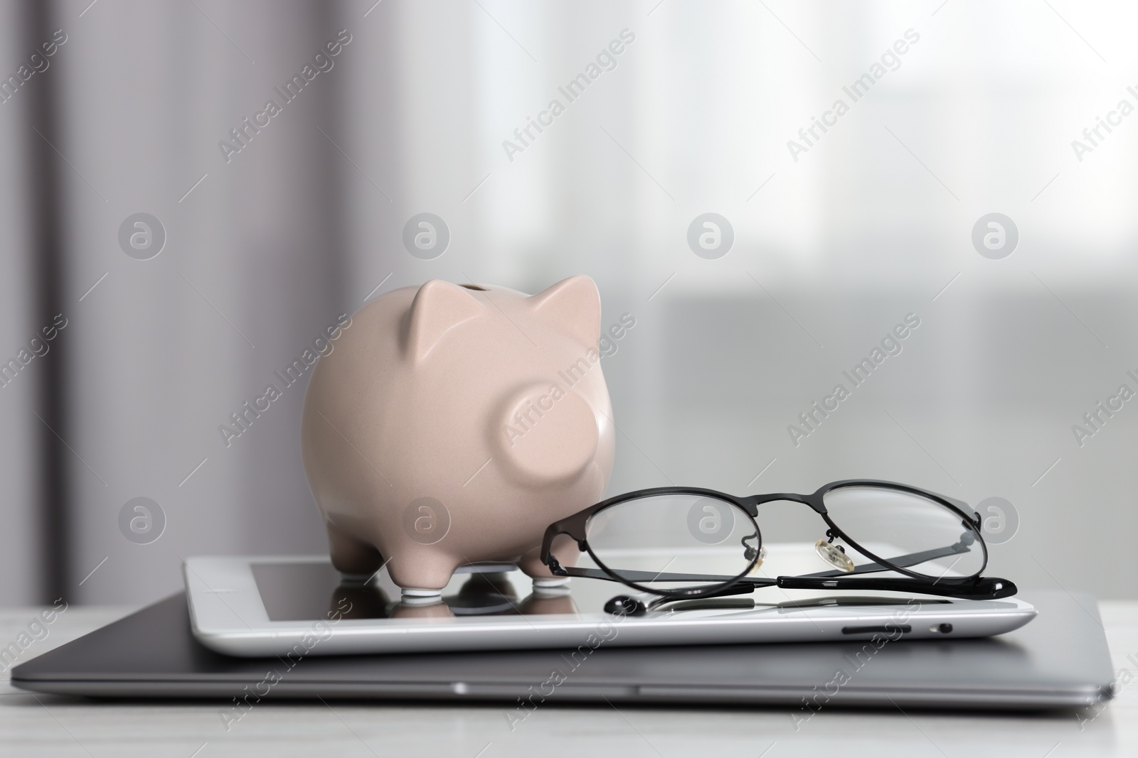 Piggy bank, glasses, tablet and laptop on white table indoors Photo of Piggy bank, glasses, tablet and laptop on white table indoors