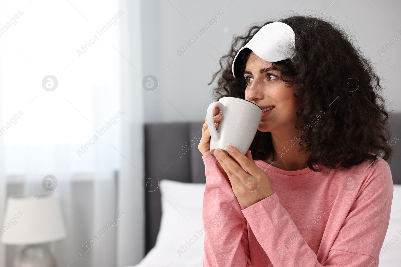 Photo of Beautiful young woman in stylish pyjama and sleep mask with cup of drink at home, space for text