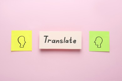 Photo of Card with word Translate between sticky notes with drawn human heads on pink background, flat lay