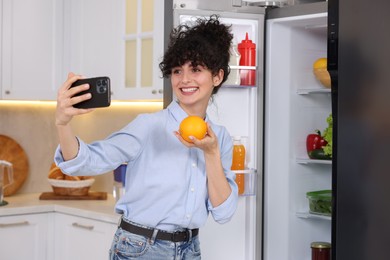 Photo of Smiling food blogger with orange taking selfie near fridge in kitchen