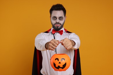 Man in scary vampire costume with fangs and pumpkin bucket on orange background. Halloween celebration