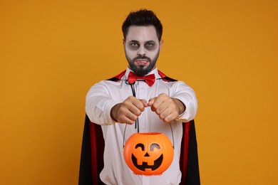 Photo of Man in scary vampire costume with fangs and pumpkin bucket on orange background. Halloween celebration