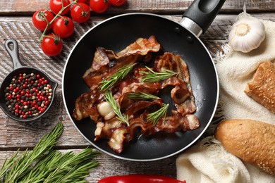 Delicious bacon slices in frying pan and products on wooden table, flat lay