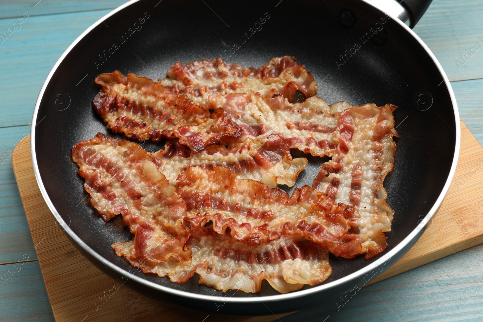 Photo of Delicious bacon slices in frying pan on blue wooden table, closeup