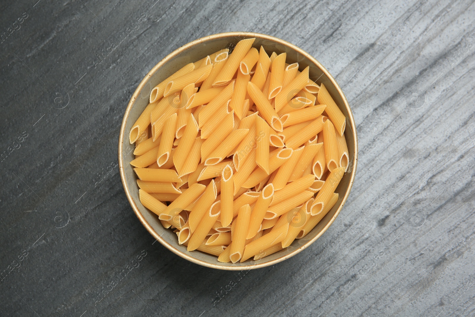 Photo of Raw penne pasta in bowl on grey table, top view