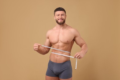 Photo of Smiling athletic man measuring waist with tape on brown background. Weight loss concept