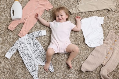 Little girl among baby clothes and detergent on carpet, top view