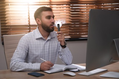 Photo of Handsome young man using disposable electronic cigarette at table in office
