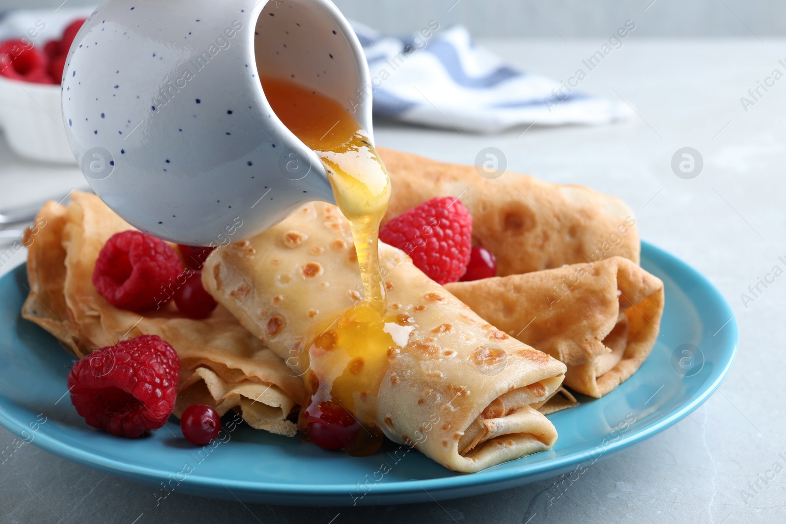 Photo of Pouring honey onto delicious crepes served with berries on light grey table, closeup