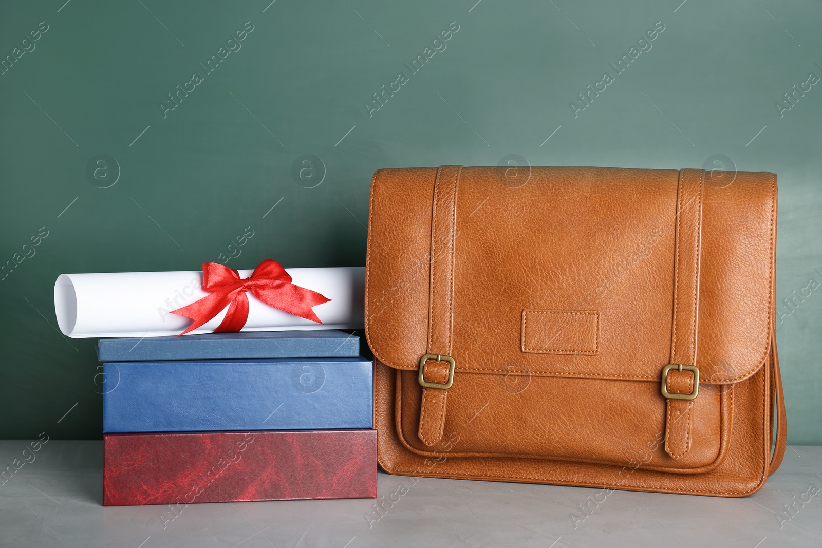 Photo of Graduate diploma with books and briefcase on table near chalkboard