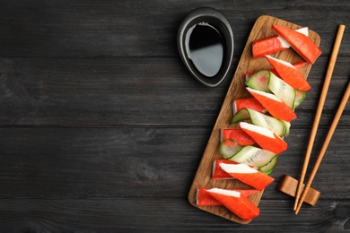 Photo of Crab sticks with cucumber and soy sauce served on dark wooden table, flat lay. Space for text
