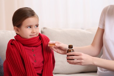 Mother giving cough syrup to her daughter on sofa indoors