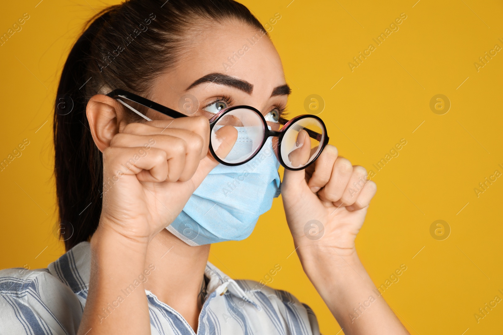 Photo of Woman wiping foggy glasses caused by wearing medical mask on yellow background, closeup