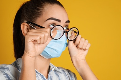 Photo of Woman wiping foggy glasses caused by wearing medical mask on yellow background, closeup