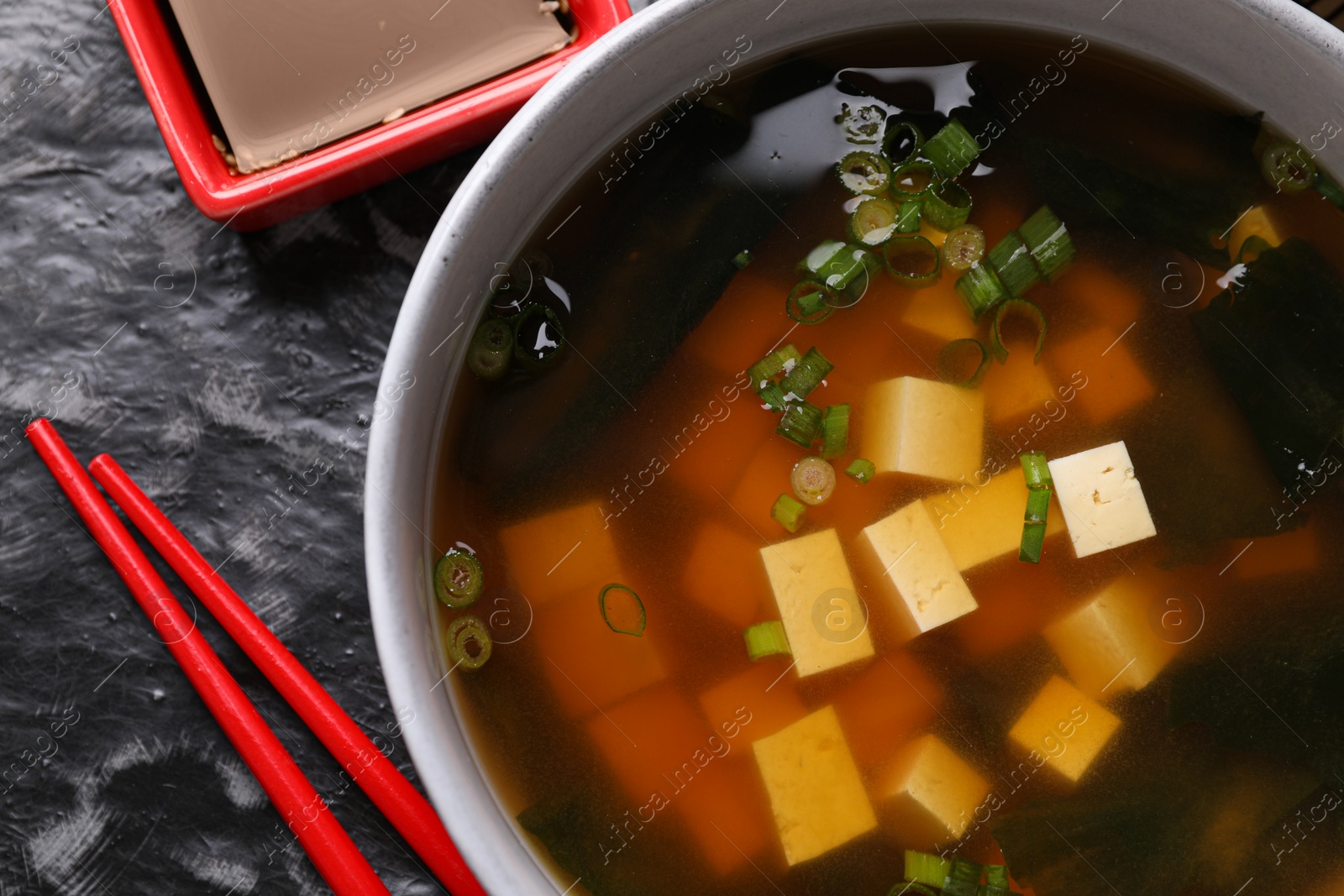 Photo of Bowl of delicious miso soup with tofu served on black textured table, flat lay