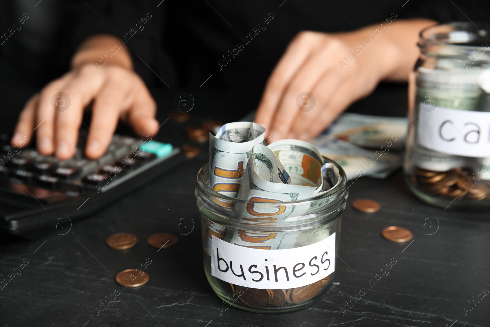 Photo of Woman using calculator near glass jar with money and tag BUSINESS on black table, closeup