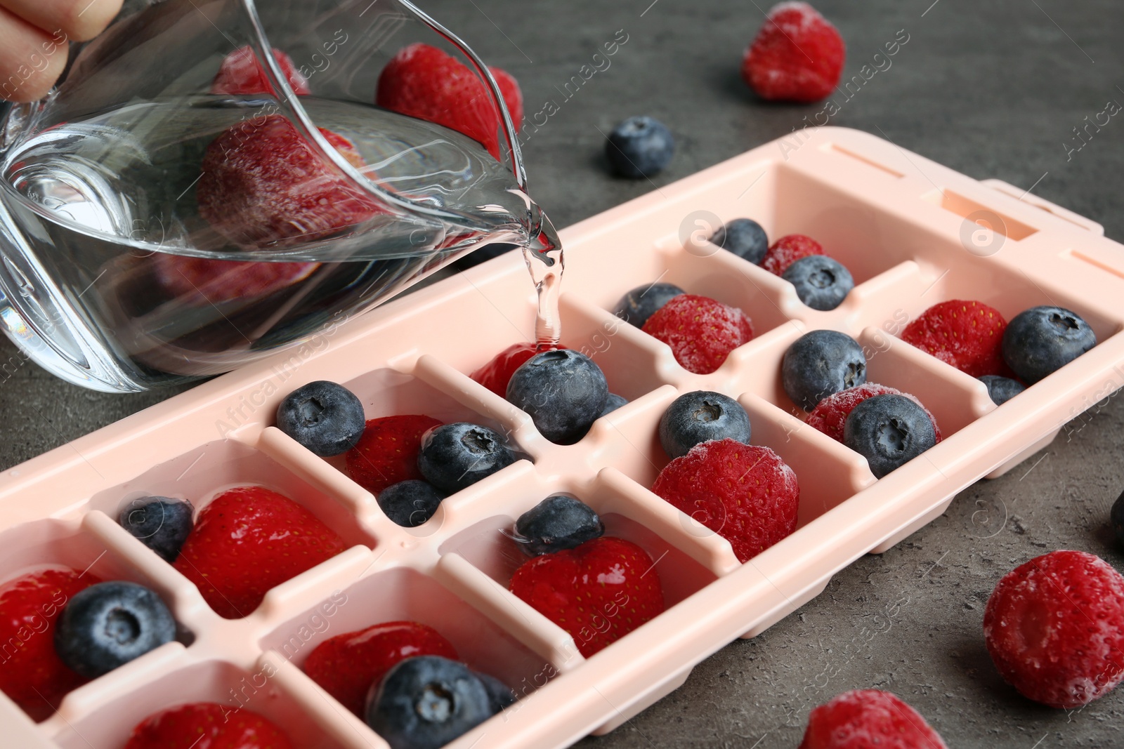 Photo of Pouring water into ice cube tray with berries on table, closeup