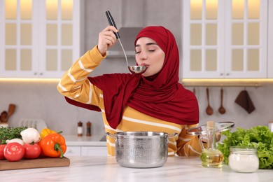 Photo of Muslim woman making delicious soup with vegetables at white table in kitchen